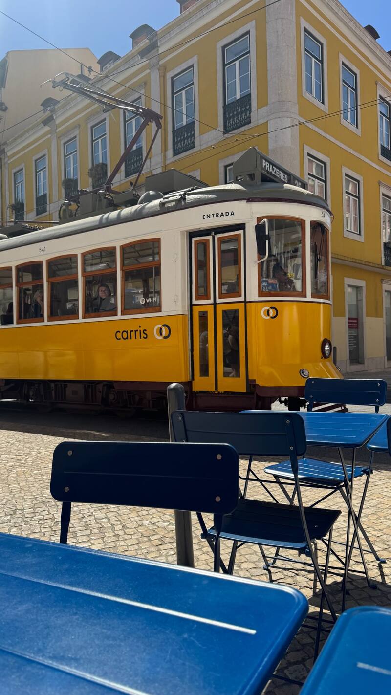 Um elétrico amarelo passa junto à esplanada, com mesas e cadeiras azuis à frente e um prédio amarelo atrás.
