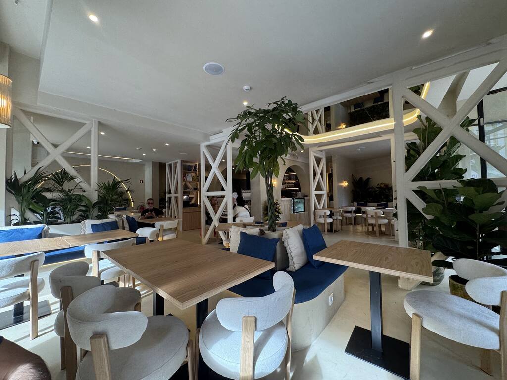 Vista larga da sala: divisórias brancas de madeira em X, banco corrido com almofadas azuis, mesas de madeira clara e cadeiras brancas.
