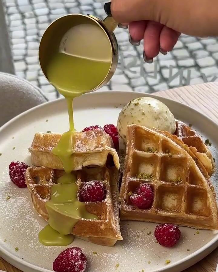 Waffles com framboesas e uma bola de gelado, com molho verde a cair de um jarrinho dourado.