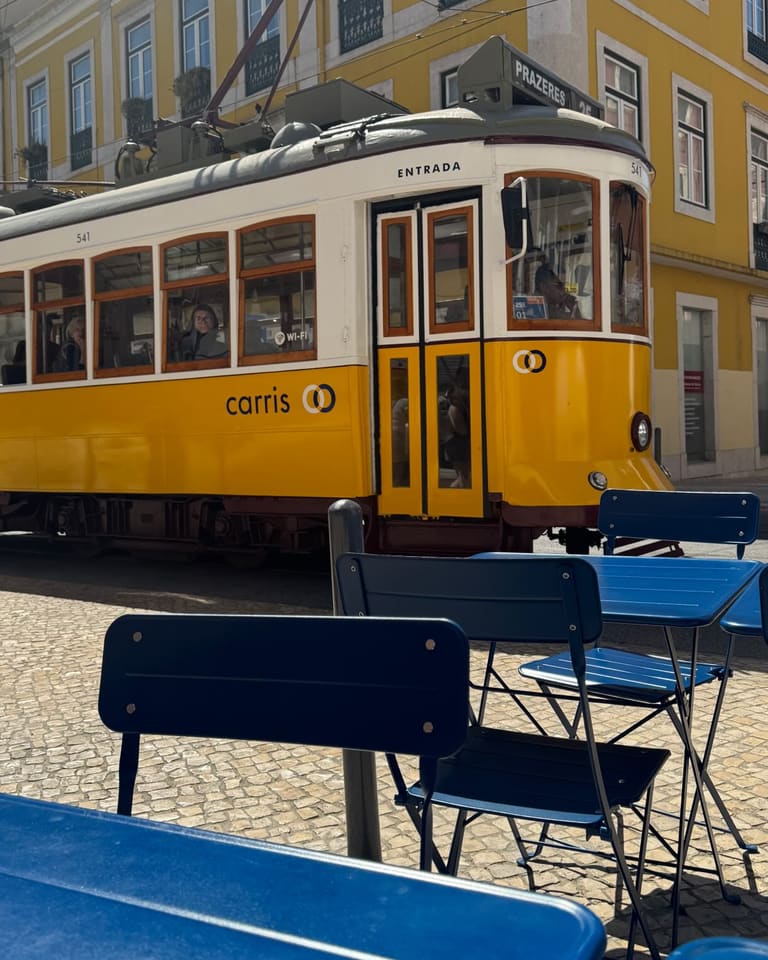 Um elétrico amarelo passa junto à esplanada, com mesas e cadeiras azuis à frente e um prédio amarelo atrás.