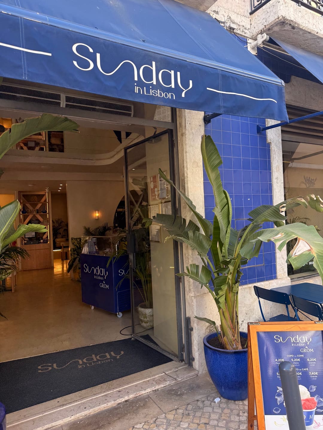 A entrada do Sunday in Lisbon: o toldo azul-marinho com o nome da casa, uma planta grande num vaso azul, a parede de azulejo azul e a porta aberta para a sala.