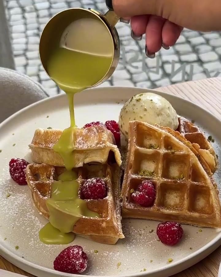 Waffles com framboesas e uma bola de gelado, com molho verde a cair de um jarrinho dourado.