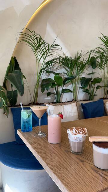 Bebidas numa mesa de madeira, com o banco azul e plantas atrás: uma bebida azul com lima, uma bebida cremosa num copo de pé, um batido cor-de-rosa com um morango, um copo coberto de natas polvilhadas e outro com cacau e um biscoito.