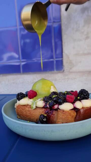 Uma French Toast com natas, amoras, framboesas e mirtilos, e molho verde a cair de um jarrinho dourado, diante de azulejo azul.
