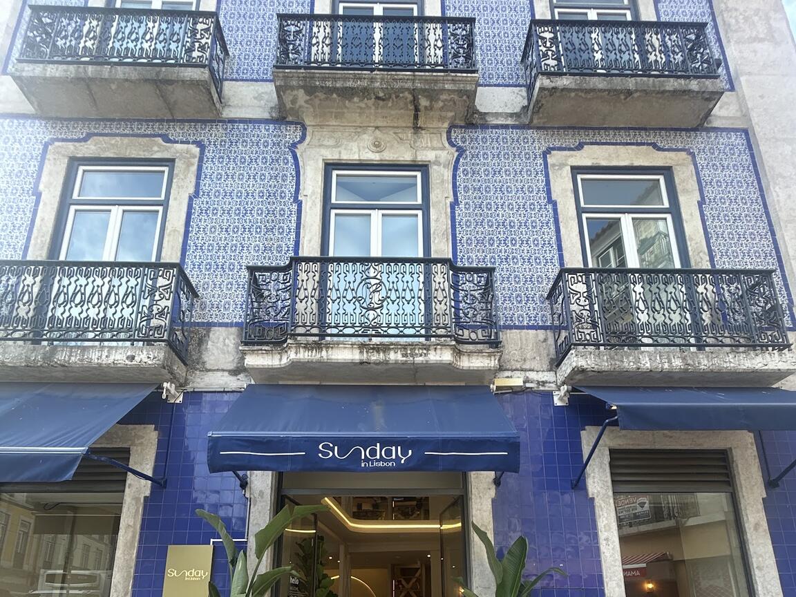 O prédio de azulejo azul, com varandas de ferro e três toldos azul-marinho. No toldo do meio, sobre a porta, o nome Sunday in Lisbon; ao lado da porta, a placa dourada.